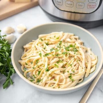 Bowl of Instant Pot Fettuccine Alfredo sitting next to Instant Pot and fresh garlic cloves.