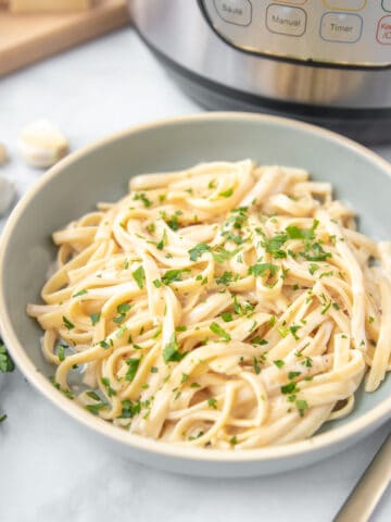 Bowl of Instant Pot Fettuccine Alfredo sitting next to Instant Pot and fresh garlic cloves.