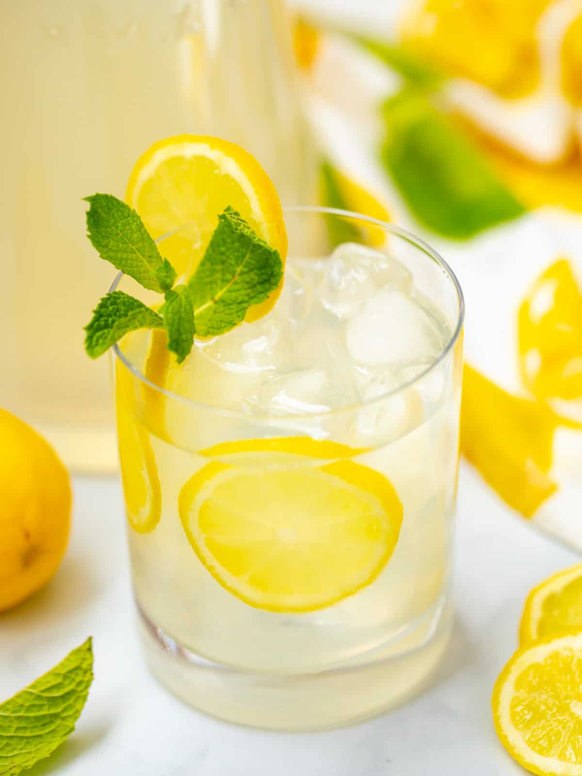 Class of mint lemonad with sliced lemon and fresh mint next to fresh lemons.