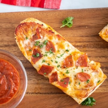 French Bread pizza with pepperoni on wooden cutting board next to pizza sauce.