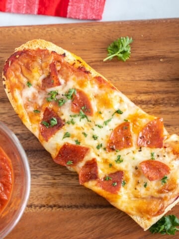 French Bread pizza with pepperoni on wooden cutting board next to pizza sauce.