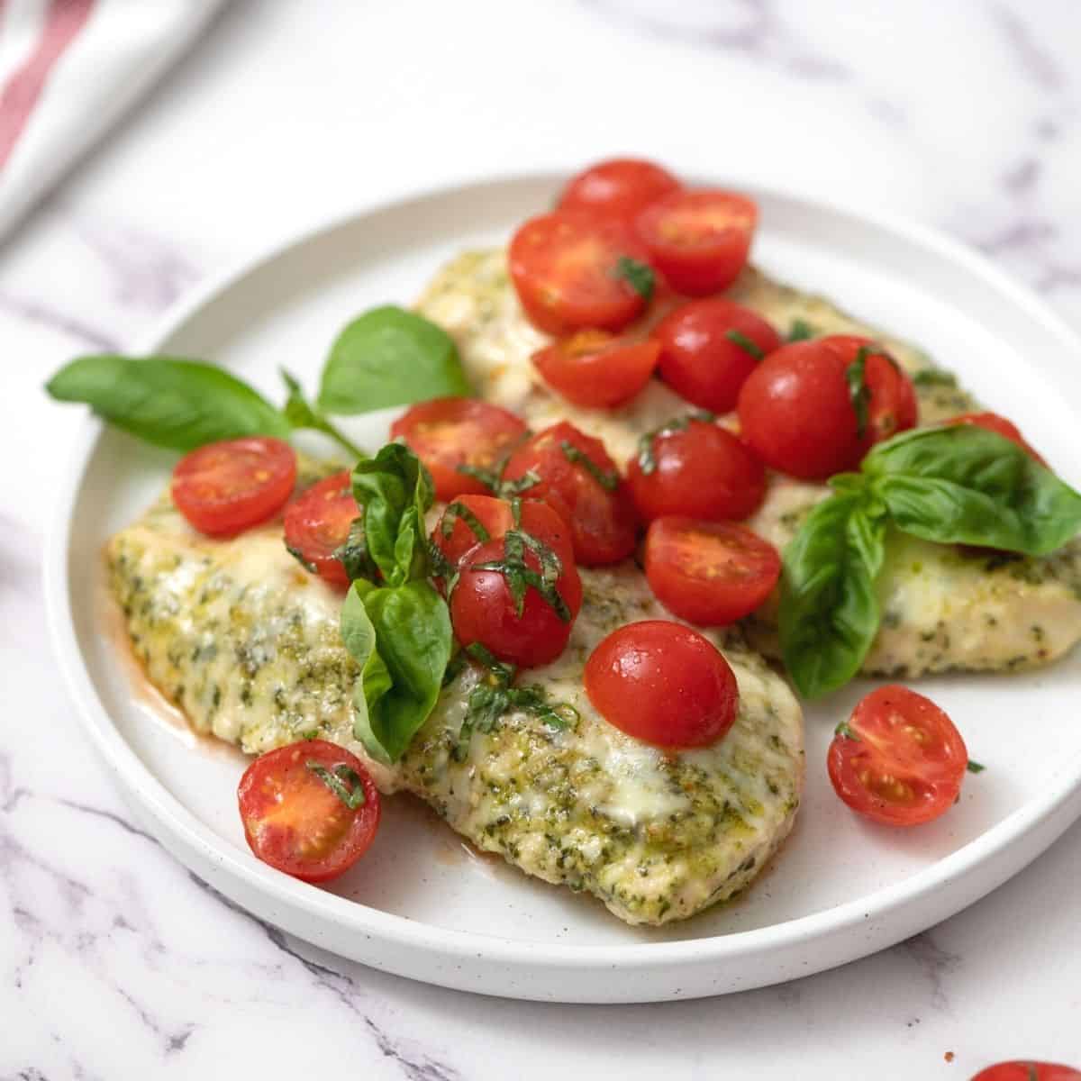 Baked Pesto Chicken topped with fresh grape tomatoes and basil on white plate. 