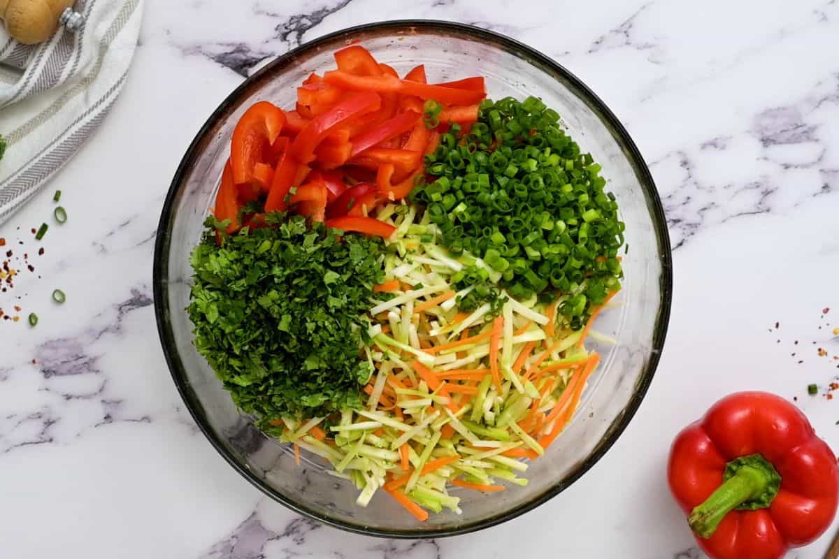Noodles with broccoli slaw, cilantro, green onions, and red pepper in glass bowl.