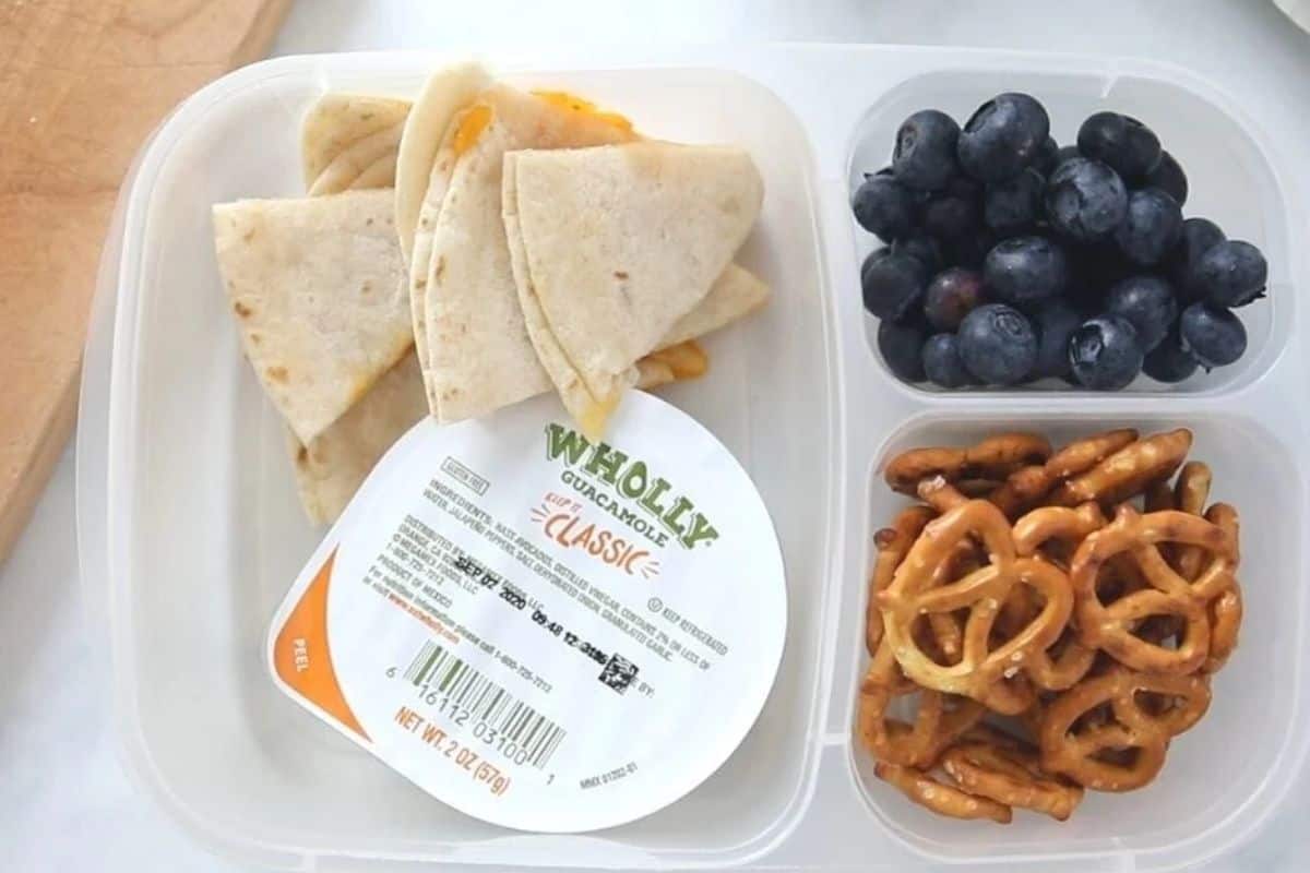 Cheese quesadilla wedges packed in bento box with guacamole cup, pretzels, and fruit.