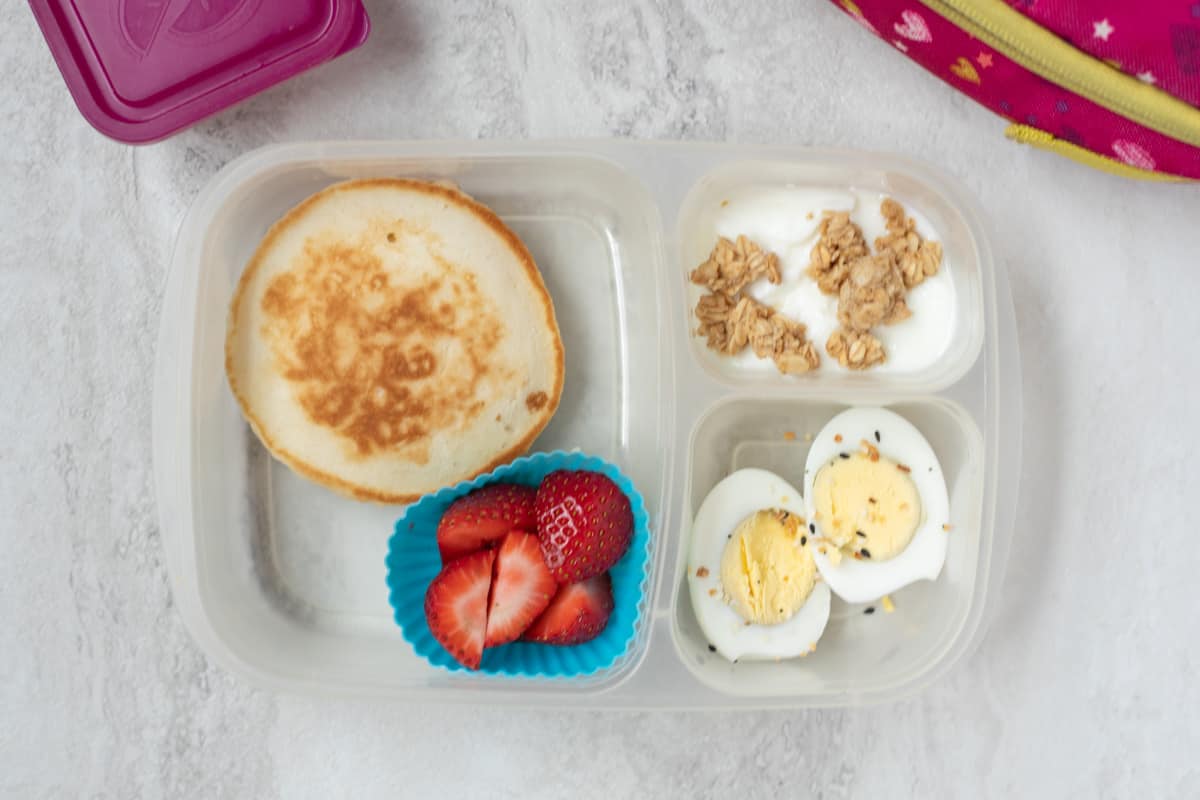 Lunch container with pancakes, syrup, strawberries, hard boiled eggs, and yogurt with granola.