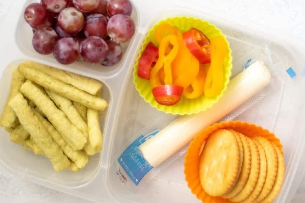Bento box with crackers, cheese stick, pea crisps, peppers, and grapes.