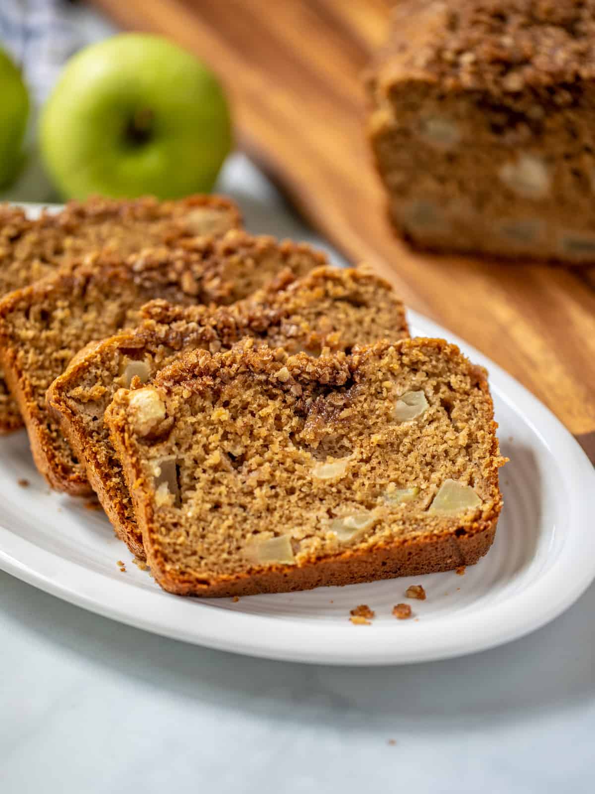 Sliced apple bread with streusel on white platter with granny smith apples in background.