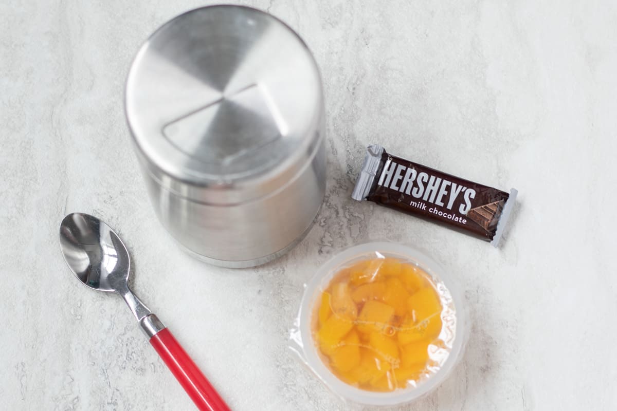 Thermos with leftovers next to spoon, fruit cup, and candy bar.