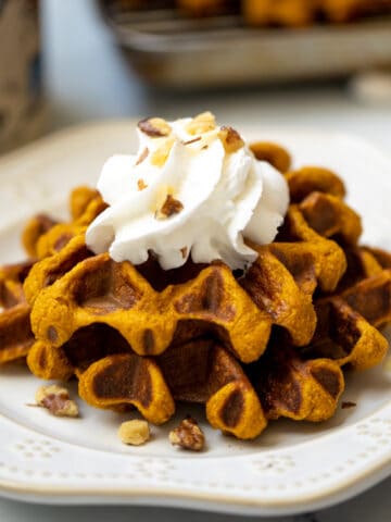 Sweet Potato waffles topped with whipped cream and crushed nuts on cream plate with maple syrup in background.