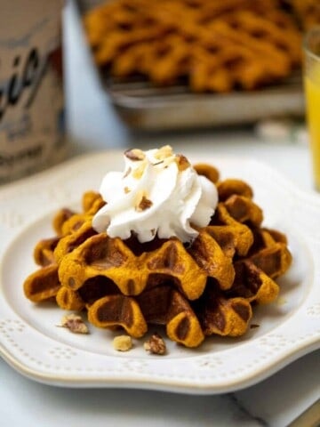 Sweet Potato waffles topped with whipped cream and crushed nuts on cream plate with maple syrup in background.