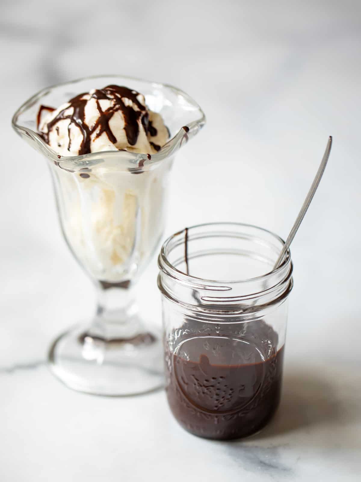 Jar of homemade chocolate syrup next to vanilla ice cream sundae.