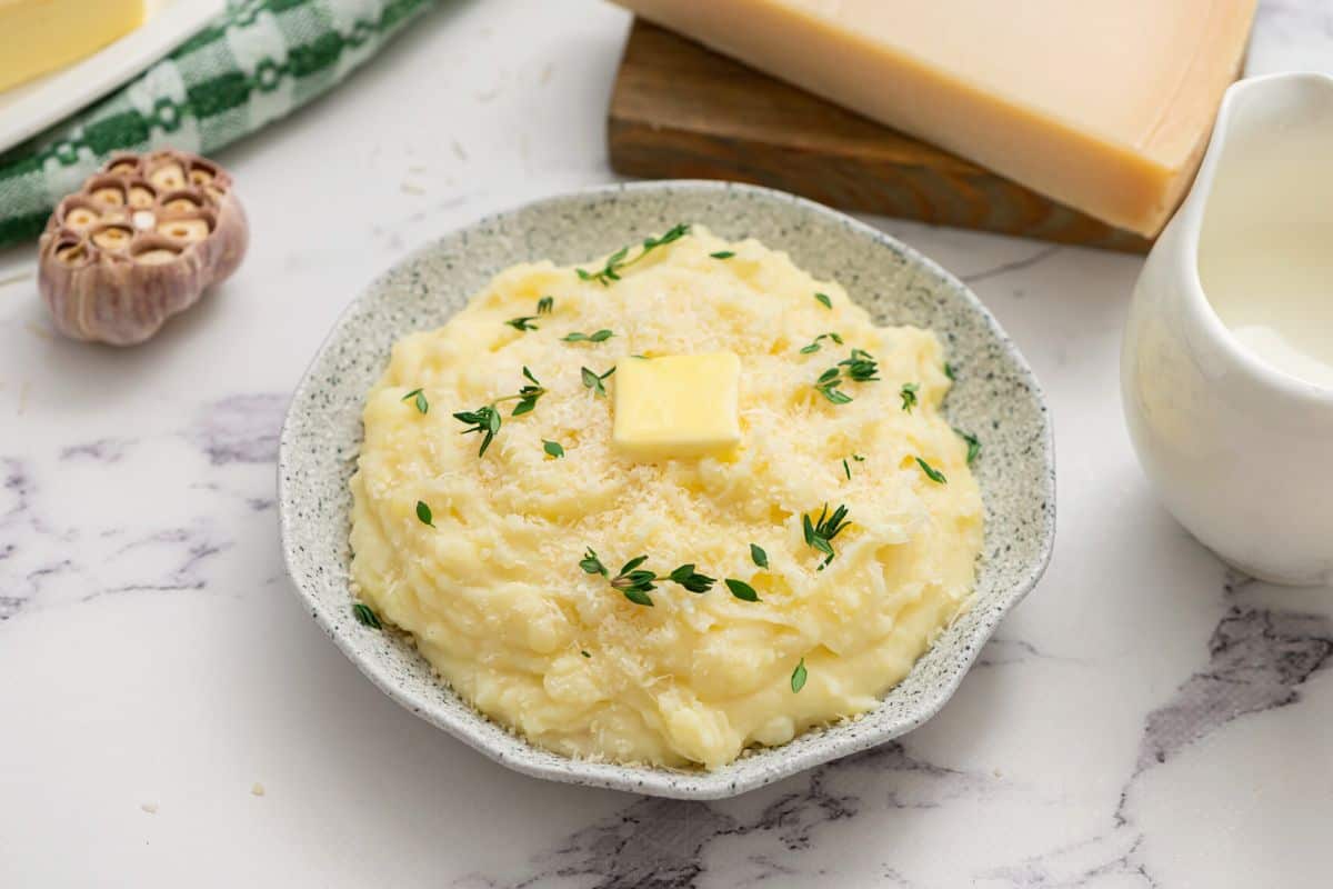 Bowl of crockpot mashed potatoes topped with butter next to cream and parmesan wedge.