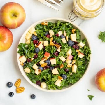 Kale salad with apples, berries and nuts in bowl with pecans and apples in background.