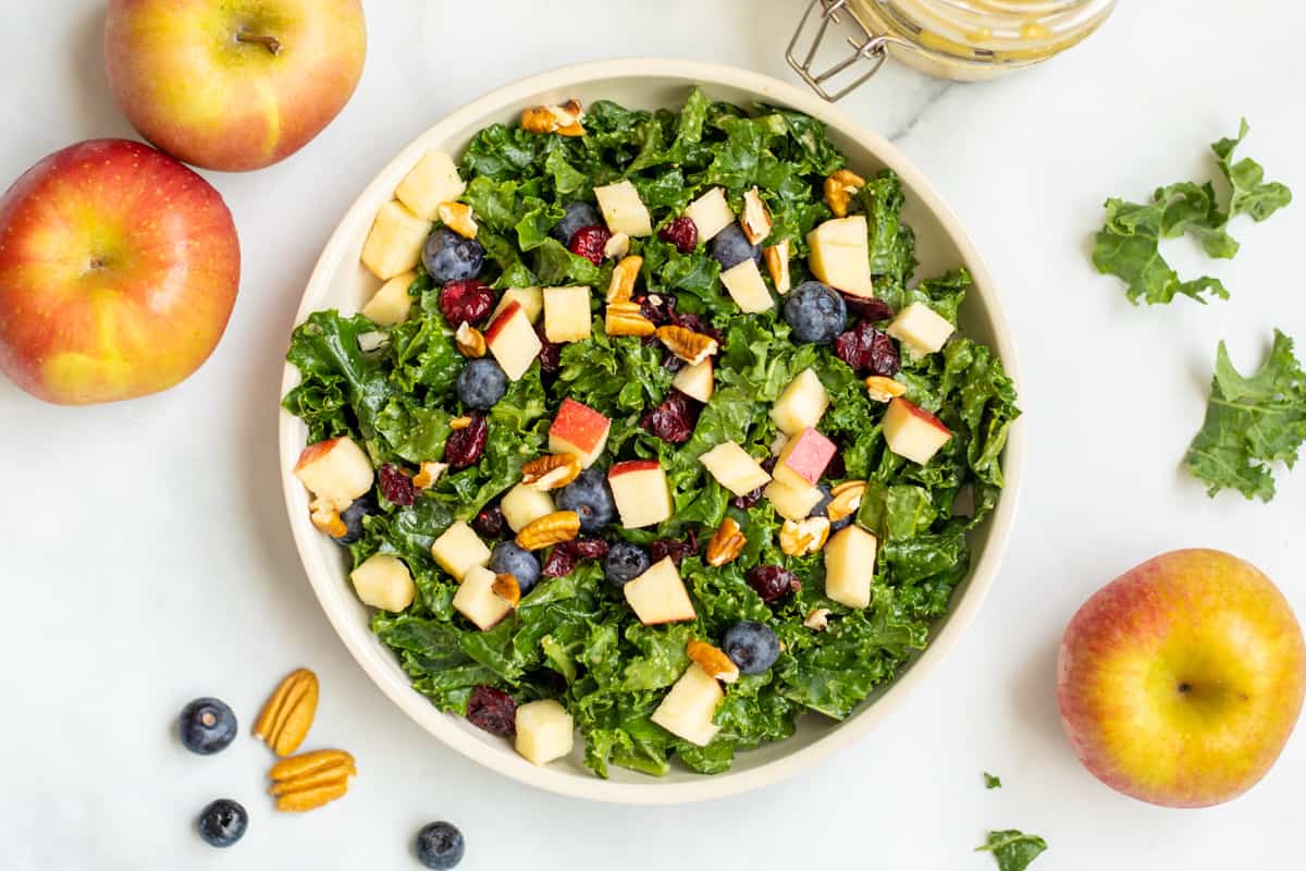Kale salad with apples, berries and nuts in bowl with pecans and apples in background.