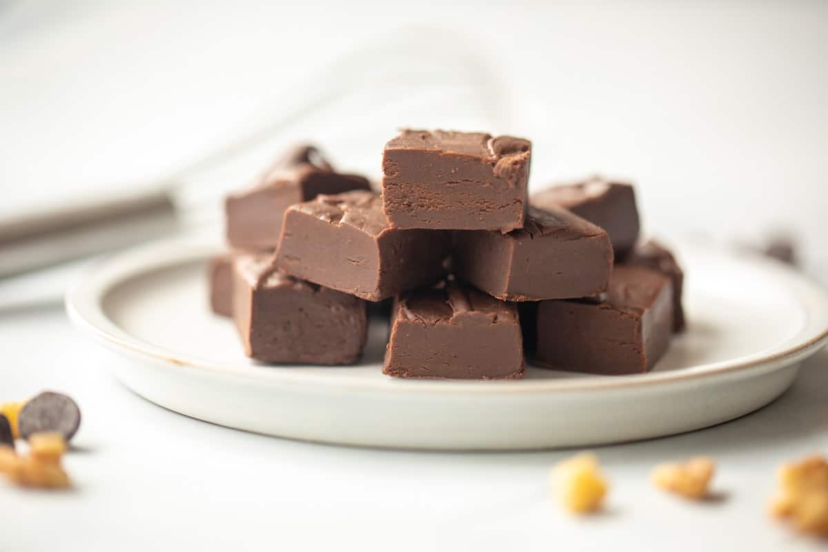 Fudge cut into cubes on white plate with nuts and chocolate chips in background.