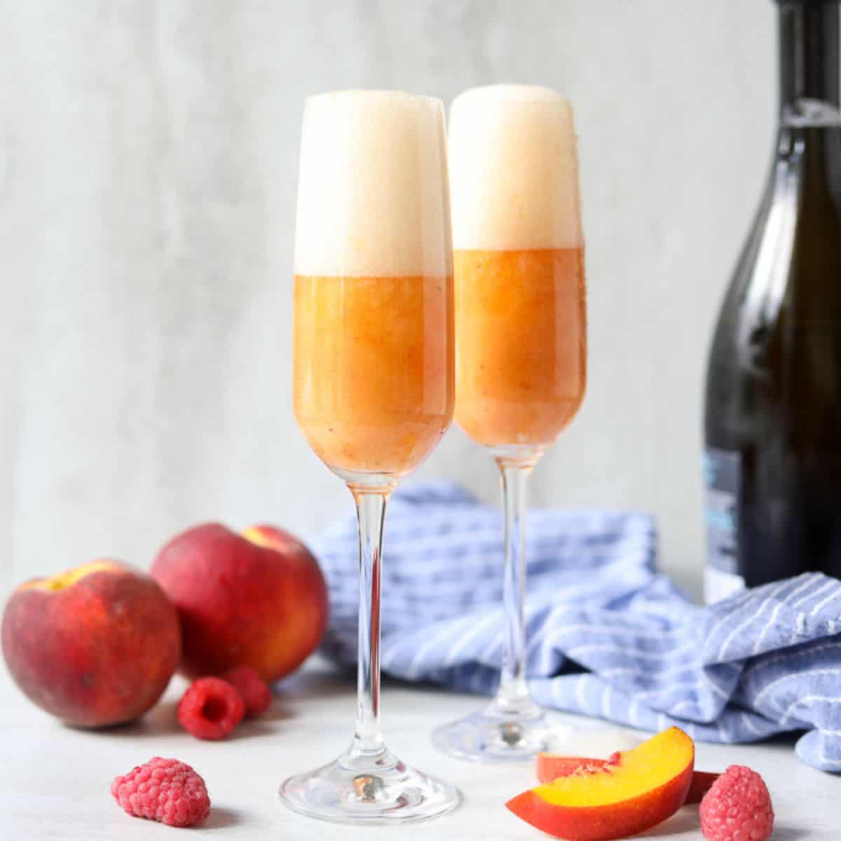 Two glasses of peach bellini cocktails next to fresh peaches and prosecco.