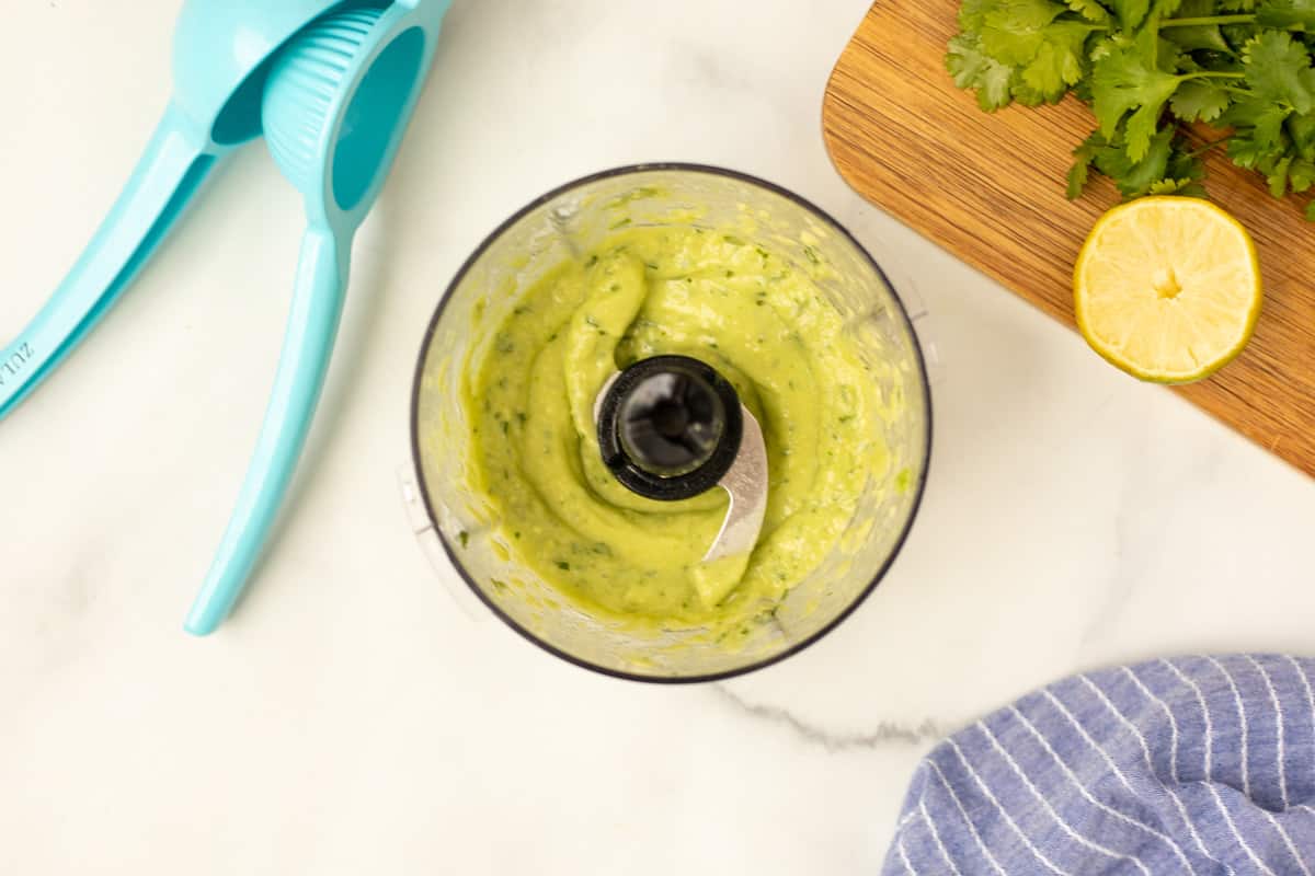 Blended avocado lime sauce in small food processor next to citrus juicer.