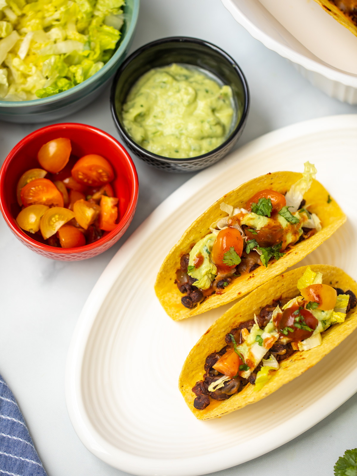 Two black bean tacos on white plate topped with diced tomatoes, lettuce, and avocado sauce.