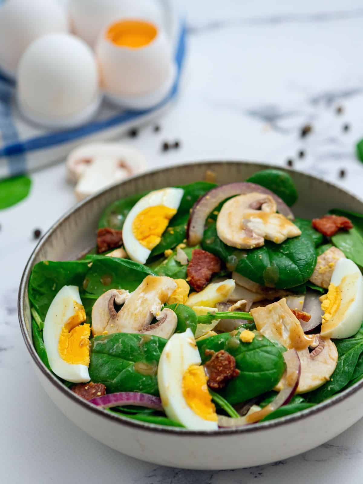 Bowl of spinach salad topped with warm bacon dressing, hard-boiled eggs, and bacon.