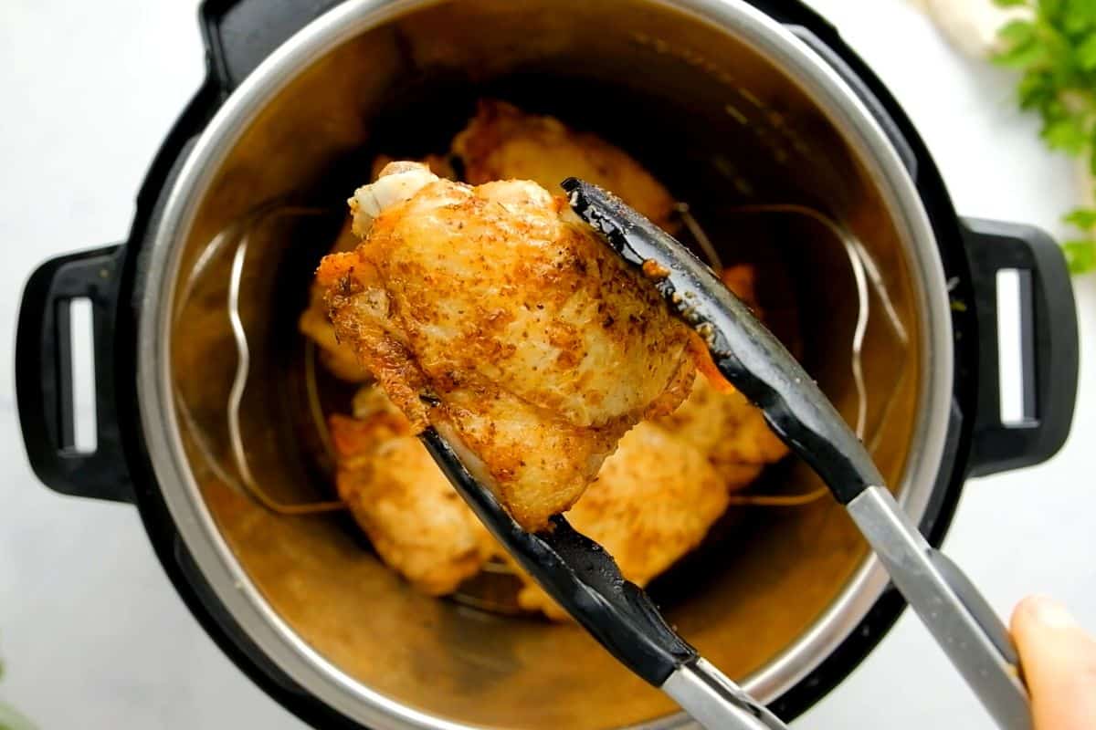 Tongs holiding crispy instant pot chicken thigh with pressure cooker in background.