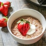Bowl of strawberries and cream oatmeal topped with strawberry and dollop of yogurt with a slow cooker in the background.
