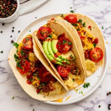 Three Assembled Breakfast tacos on white plate topped with scrambled eggs, bacon, cheese, avocado, hot sauce, and tomatoes.