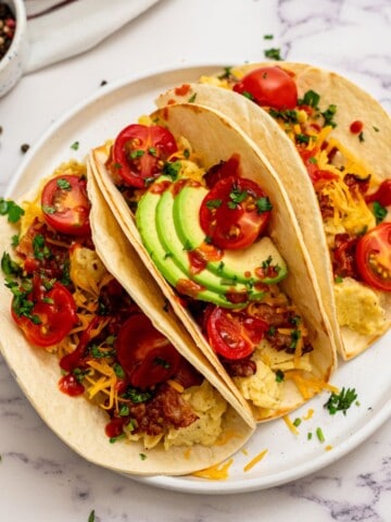 Three Assembled Breakfast tacos on white plate topped with scrambled eggs, bacon, cheese, avocado, hot sauce, and tomatoes.