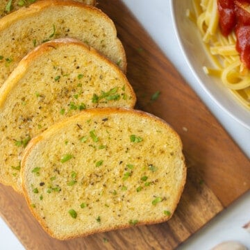 Homemade Garlic Toast on wooden cutting board with pasta in background.