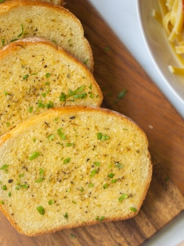 Homemade Garlic Toast on wooden cutting board with pasta in background.