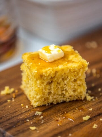Slice of cornbread topped with a pat of butter and drizzle of honey on cutting board.
