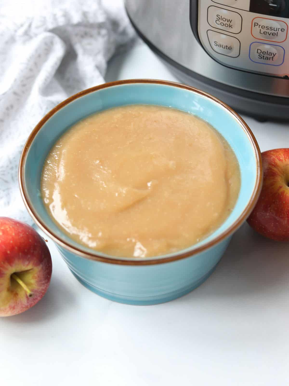 Instant Pot applesauce in blue bowl next to pressure cooker with apples in the background.