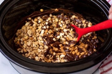 Red spoon stirring nuts into melted chocolate chips in slow cooker.