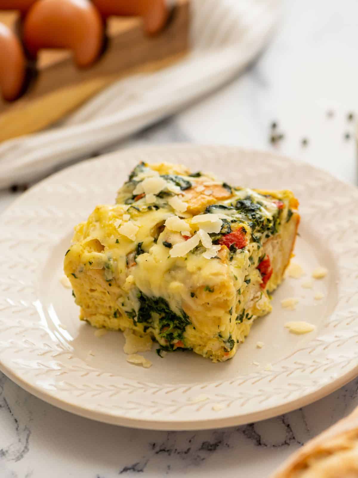 Slice of spinach and cheese breakfast strata on white plate topped with shredded parmesan cheese.