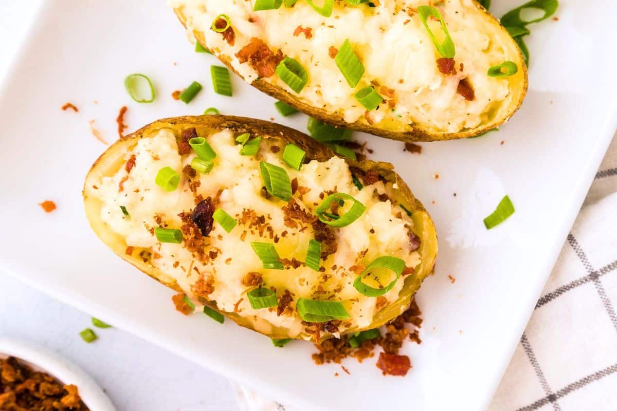 Twice baked potato topped with white cheddar, bacon, and green onions on white plate.