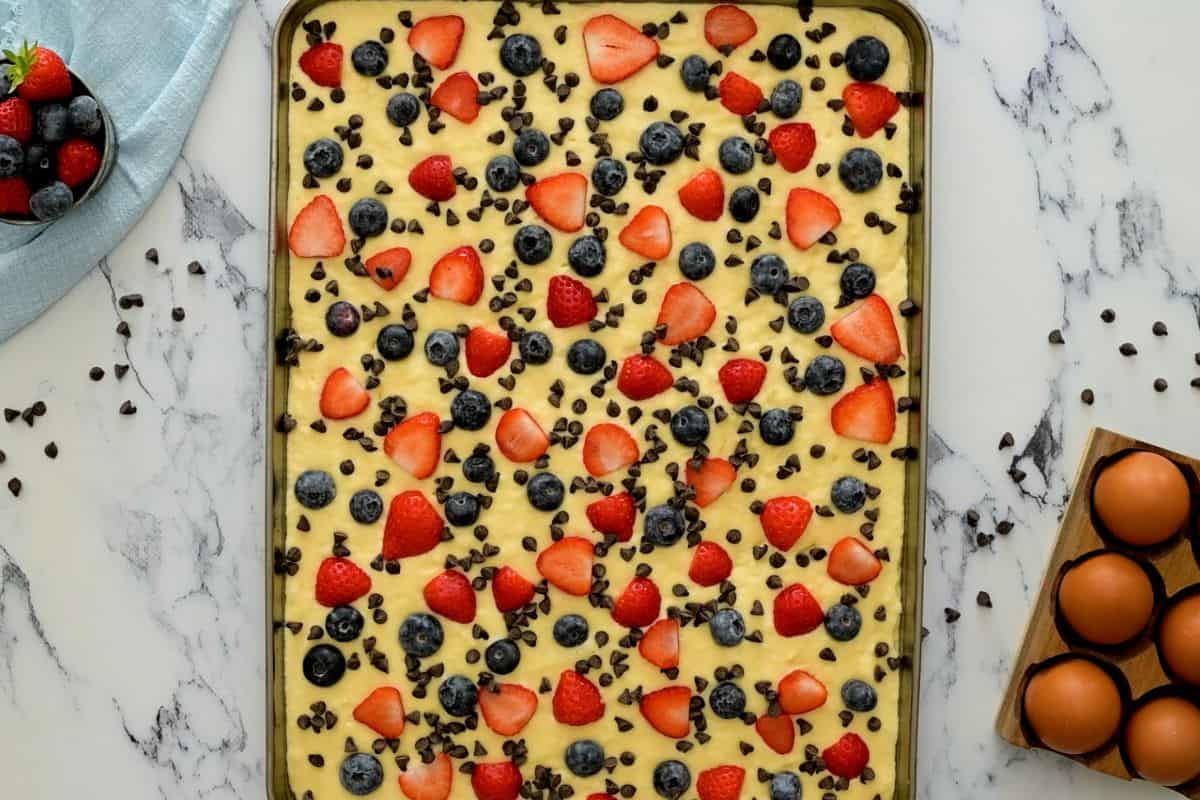 Pancake batter spread out on half-sheet pan topped with berries and mini chocolate chips.