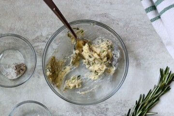 compound butter with fresh herbs mixed up with fork in small bowl next to fresh rosemary.