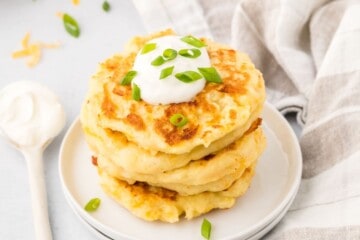 Stack of golden mashed potato cakes on plate topped with sour cream and green onions with sour cream and tray of potato cakes in background.