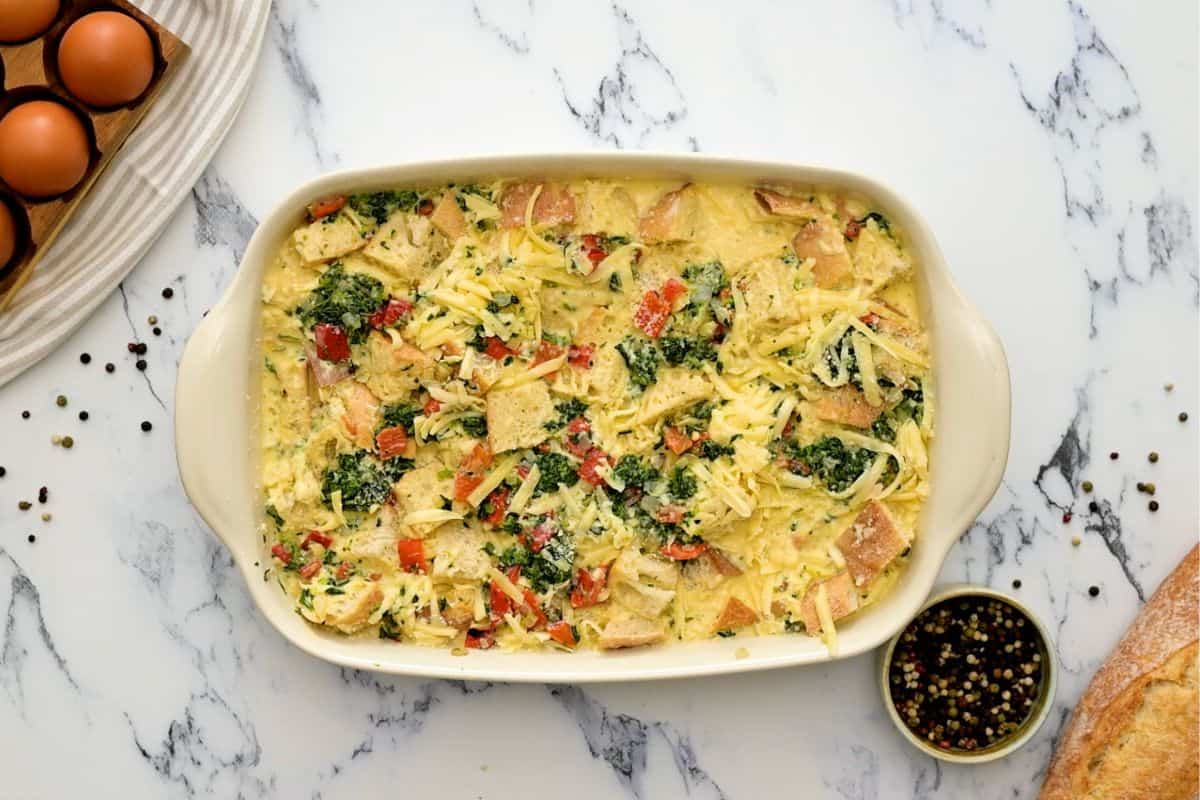 Bread, eggs, spinach, and roasted red peppers in baking dish before baking breakfast strata.