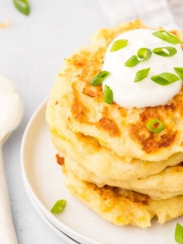 Stack of golden mashed potato cakes on plate topped with sour cream and green onions.