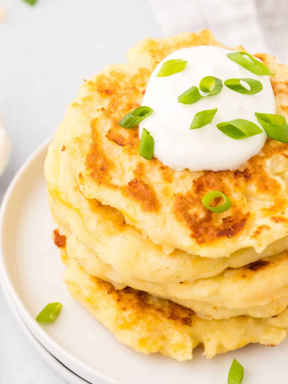 Stack of golden mashed potato cakes on plate topped with sour cream and green onions.