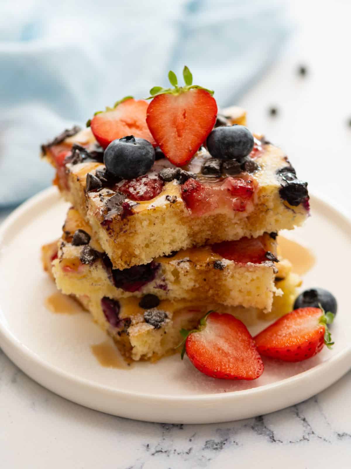 Squares of sheet pan pancakes on white plate topped with maple syrup and powdered sugar with fresh berries to the side.