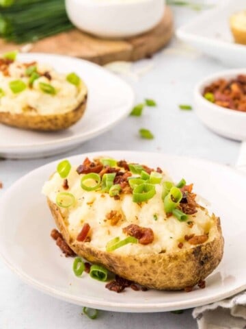 Twice baked potato topped with white cheddar, bacon, and green onions on white plate with additional double baked potatoes in background.