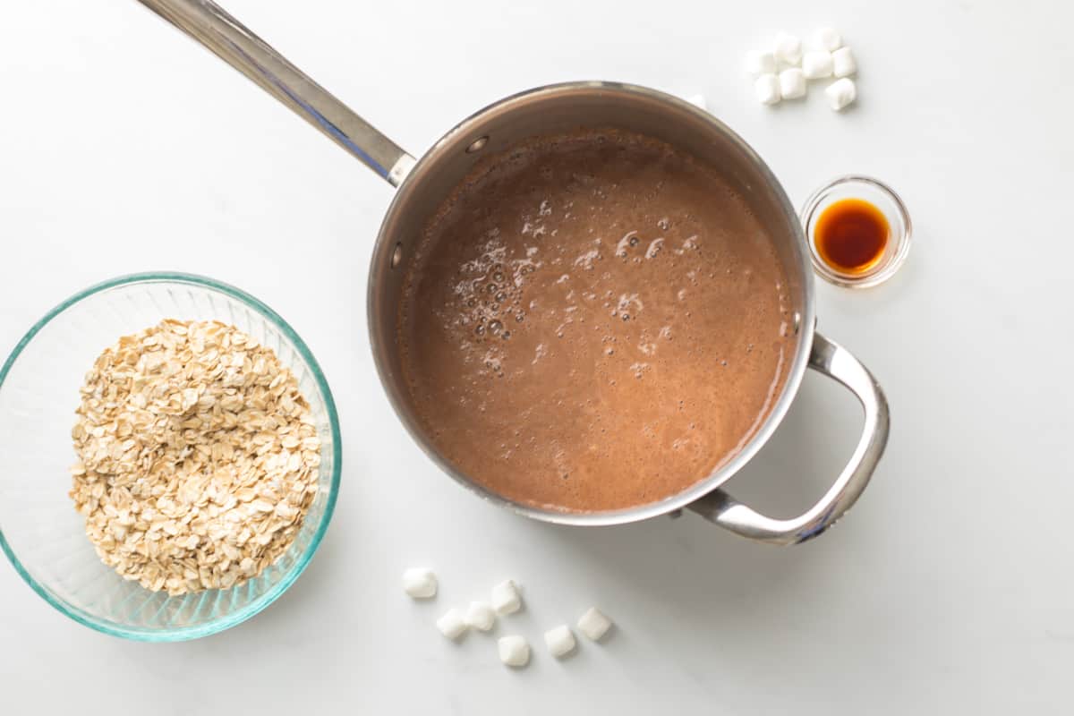 Milk, water, and cocoa powder simmering in small saucepan next to oats and marshmallows on the side.