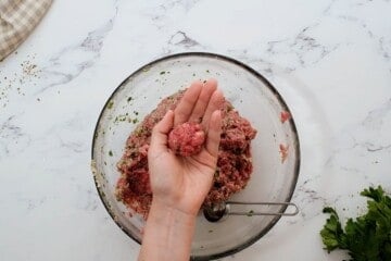 Hand showing rolled meatball with tablespoon measuring spoon in bowl with meatball mixture.