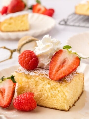 Slice of hot milk cake on white plate dusted with powdered sugar, raspberries, strawberries, and whipped cream.