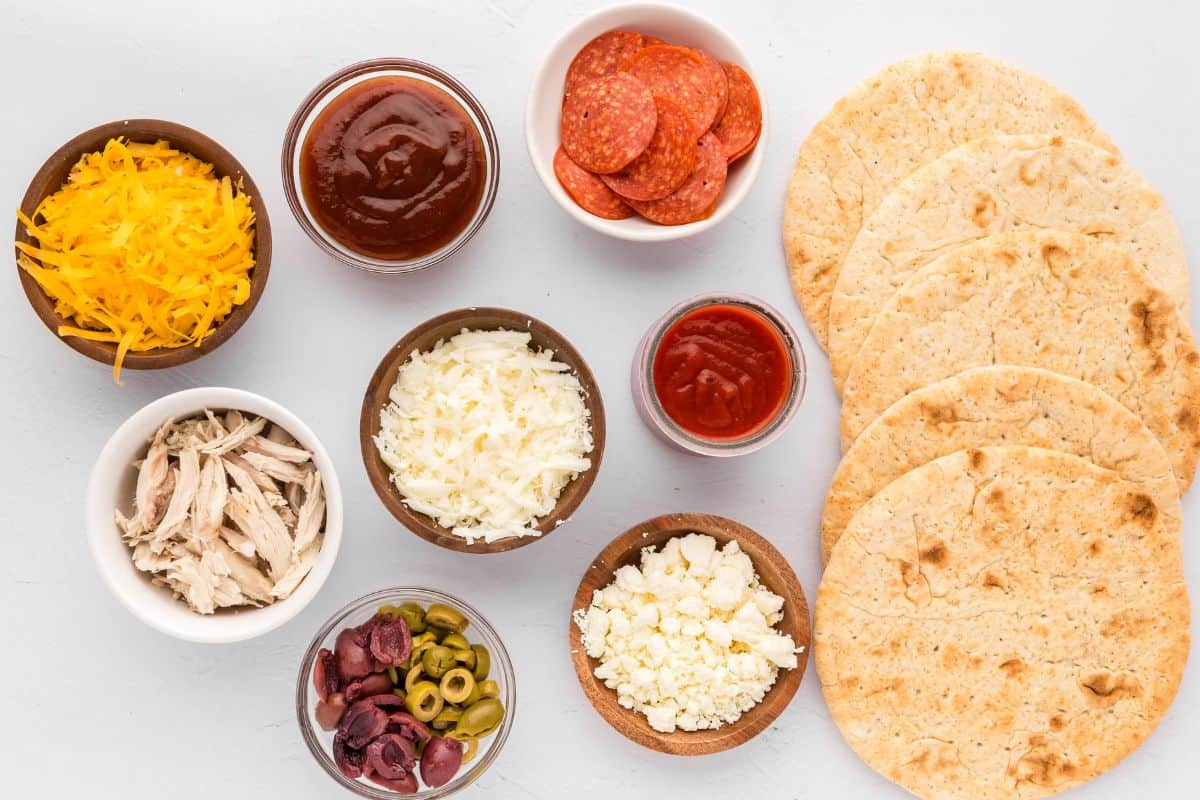 Ingredients for a variety of pita bread pizzas on counter.