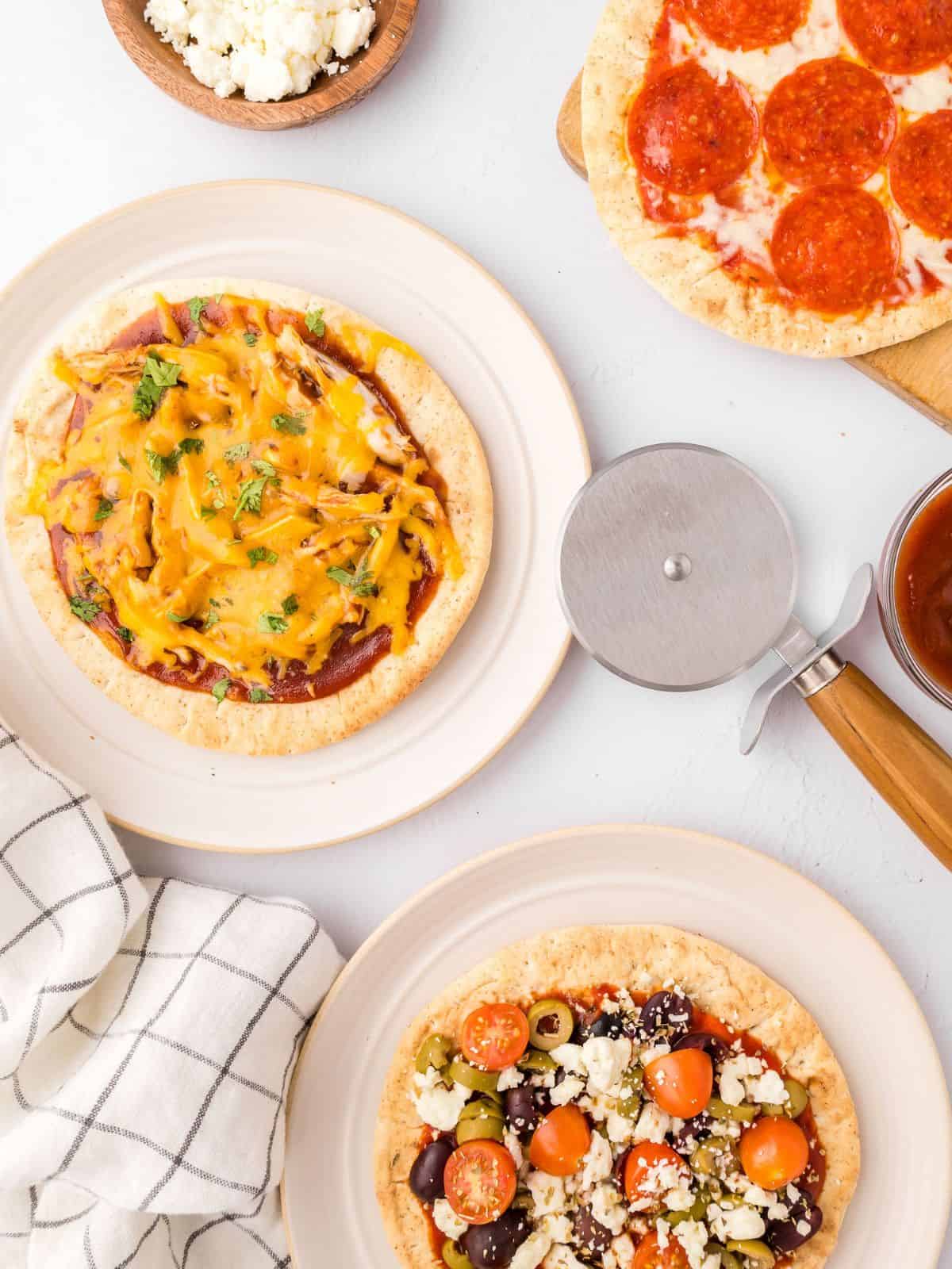 A variety of assembled and baked pita pizzas on white plates next to pizza cutter and fresh parsley.