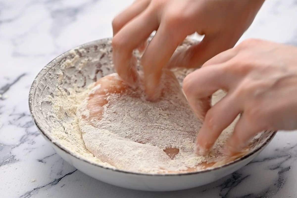 Hands dredging chicken breasts in flour.