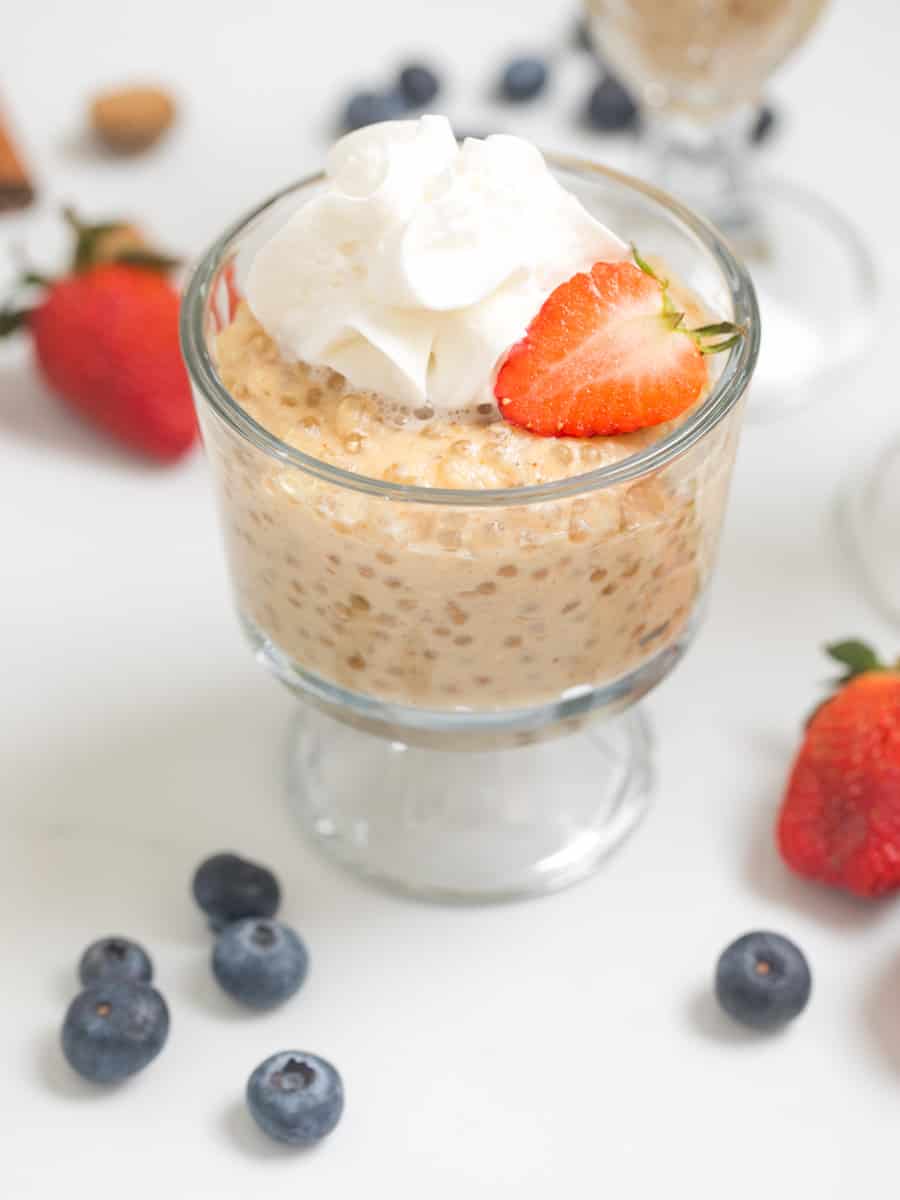Tapioca pudding in small serving bowl topped with fresh whipped cream and strawberries with berries in background.