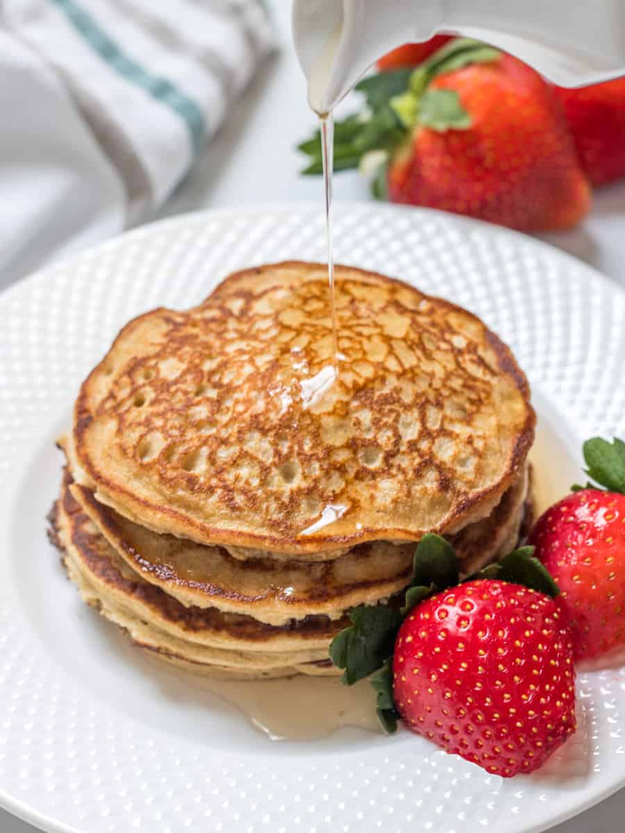 Cottage cheese pancakes stacked on white plate with syrup being poured over them next to fresh strawberries.